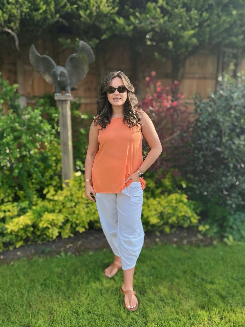 Woman in an orange vest top and white cotton pants standing in a garden