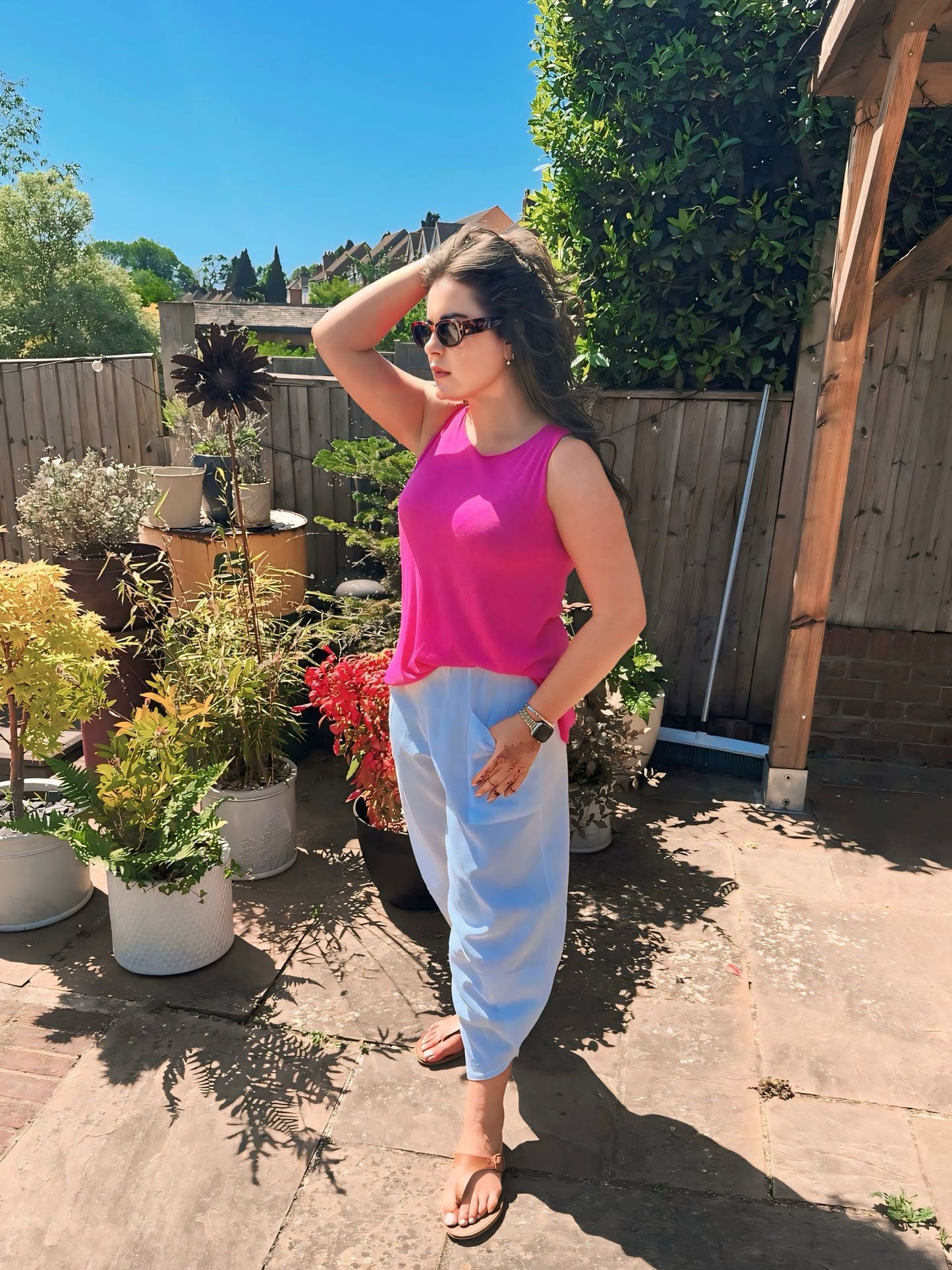 Woman in a pink top and light blue pants standing in a garden with potted plants.