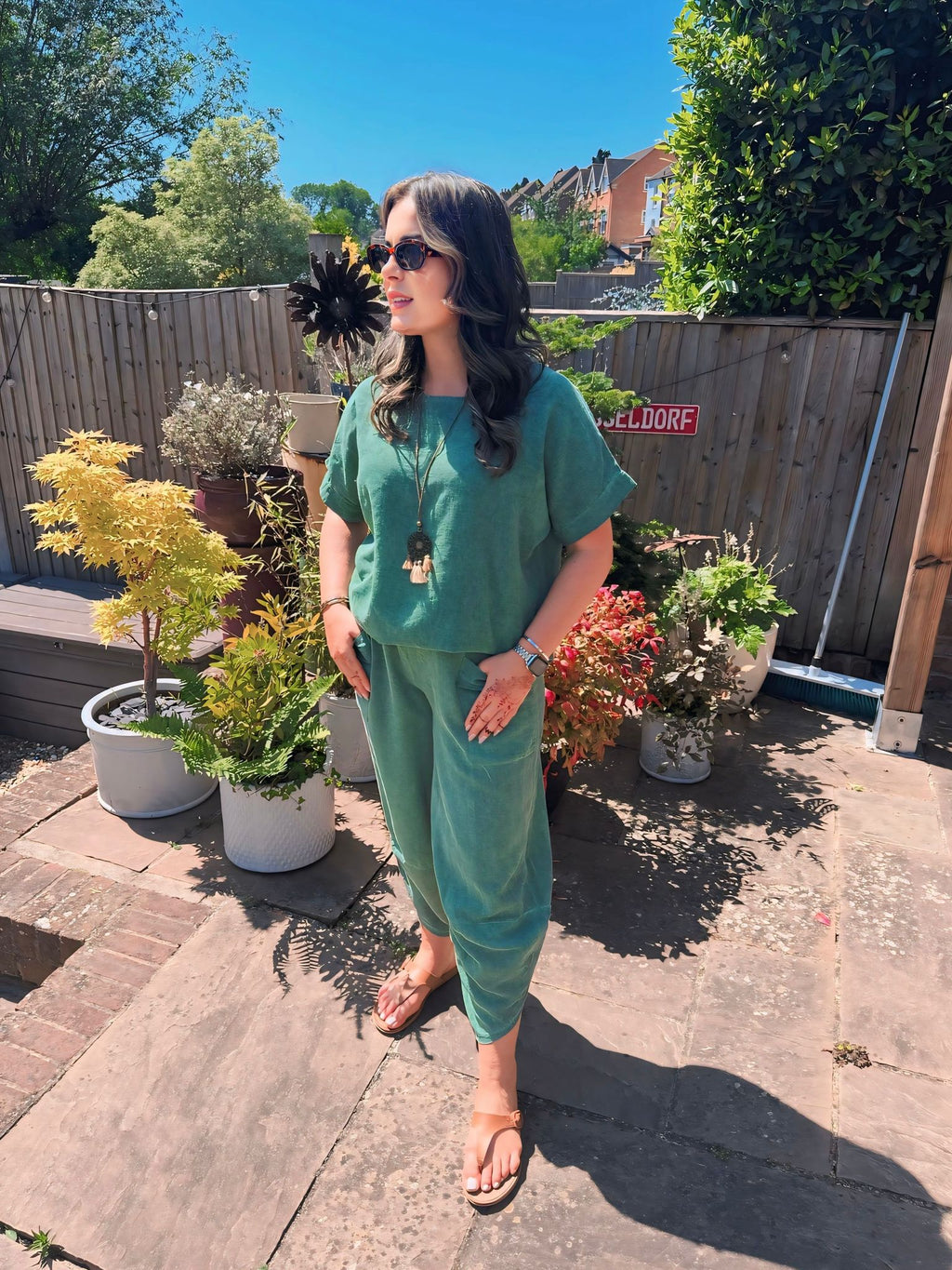 Woman in a green co-ord outfit , cotton top and cotton harem pants standing in a garden with plants and a wooden fence.