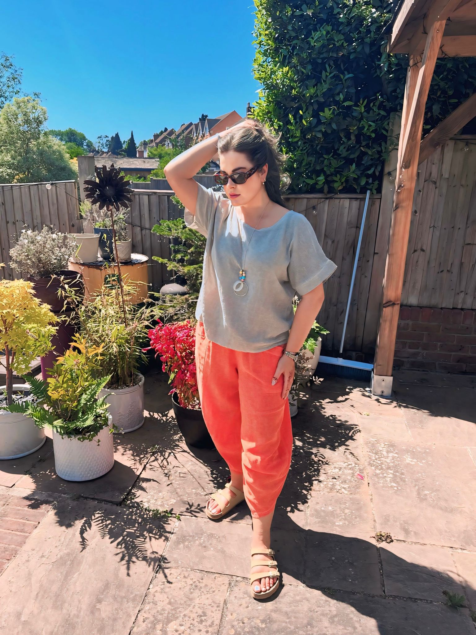 Woman standing in a garden with potted plants and a wooden fence in the background -wearing cotton top and orange cotton harem pants