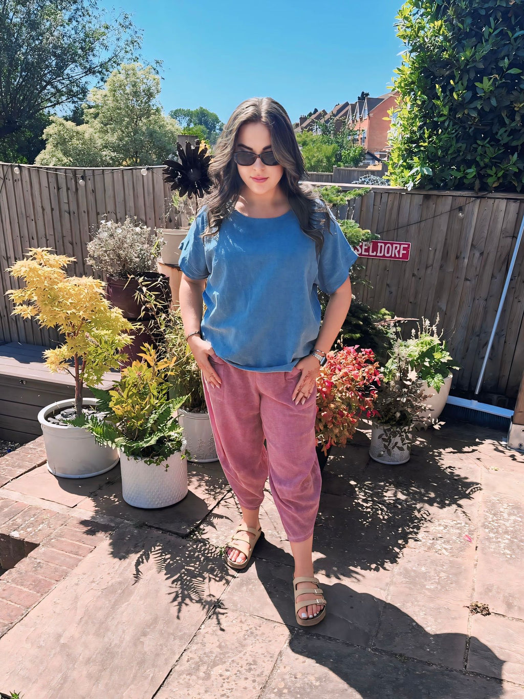 Woman standing in a garden with potted plants on a sunny day wearing a blue cotton top and dusky pink cotton harem pants